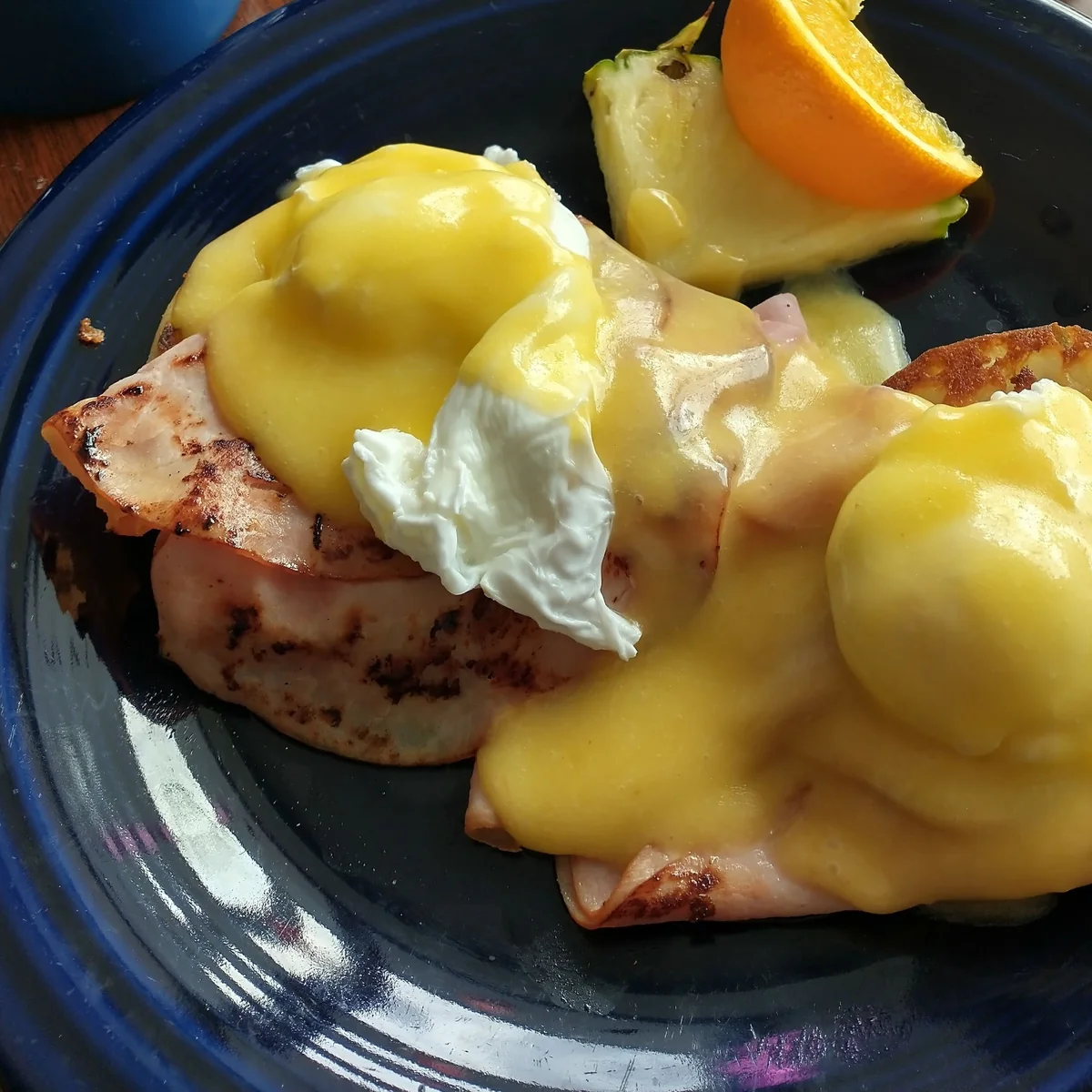 Cherie Inn eggs Benedict — two poached eggs and shaved ham under scratch hollandaise on a cobalt-blue plate, with hash browns and an orange wedge.