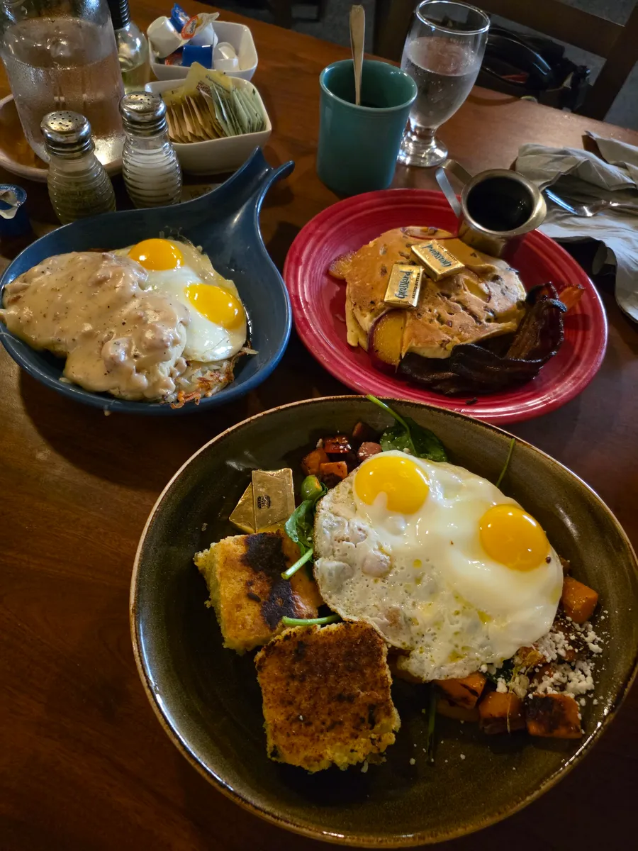 Three plates on a table: biscuits and gravy with eggs, pancakes with bacon, and a veggie hash bowl